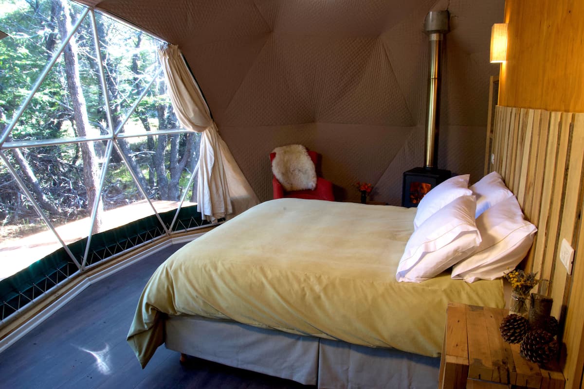 A cozy room with a bed, a window with curtains, a wooden table with a vase, and a wood-burning stove.