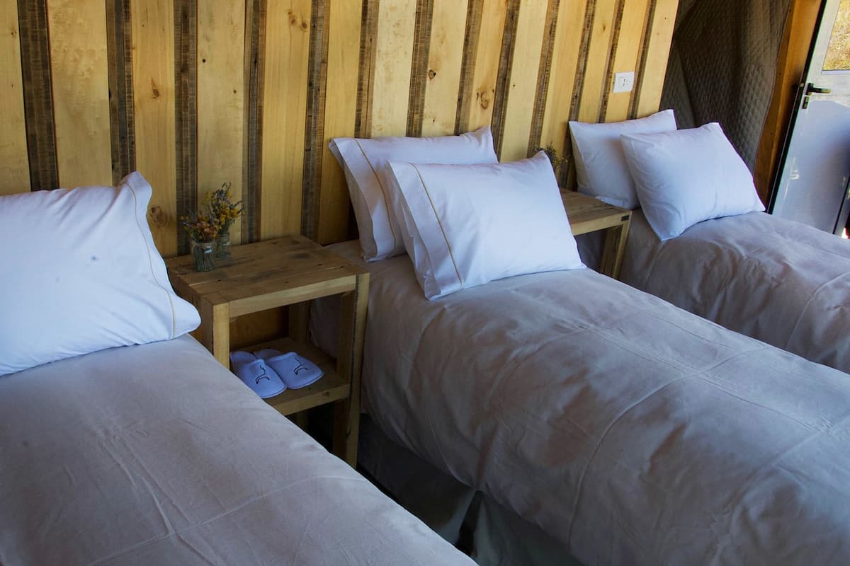 A room with two beds, each with white bedding and pillows, and a wooden nightstand with a vase of flowers and slippers.