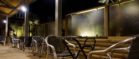 Un espacio al aire libre con sillas y mesas bajo un techo de madera, con una pared de diseño texturizado y una planta de palma.