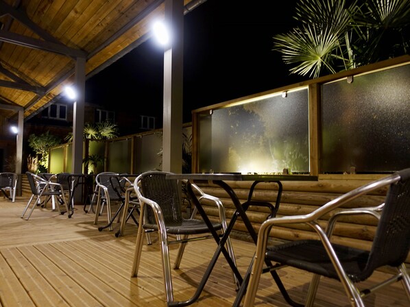 Un espacio al aire libre con sillas y mesas bajo un techo de madera, con una pared de diseño texturizado y una planta de palma.