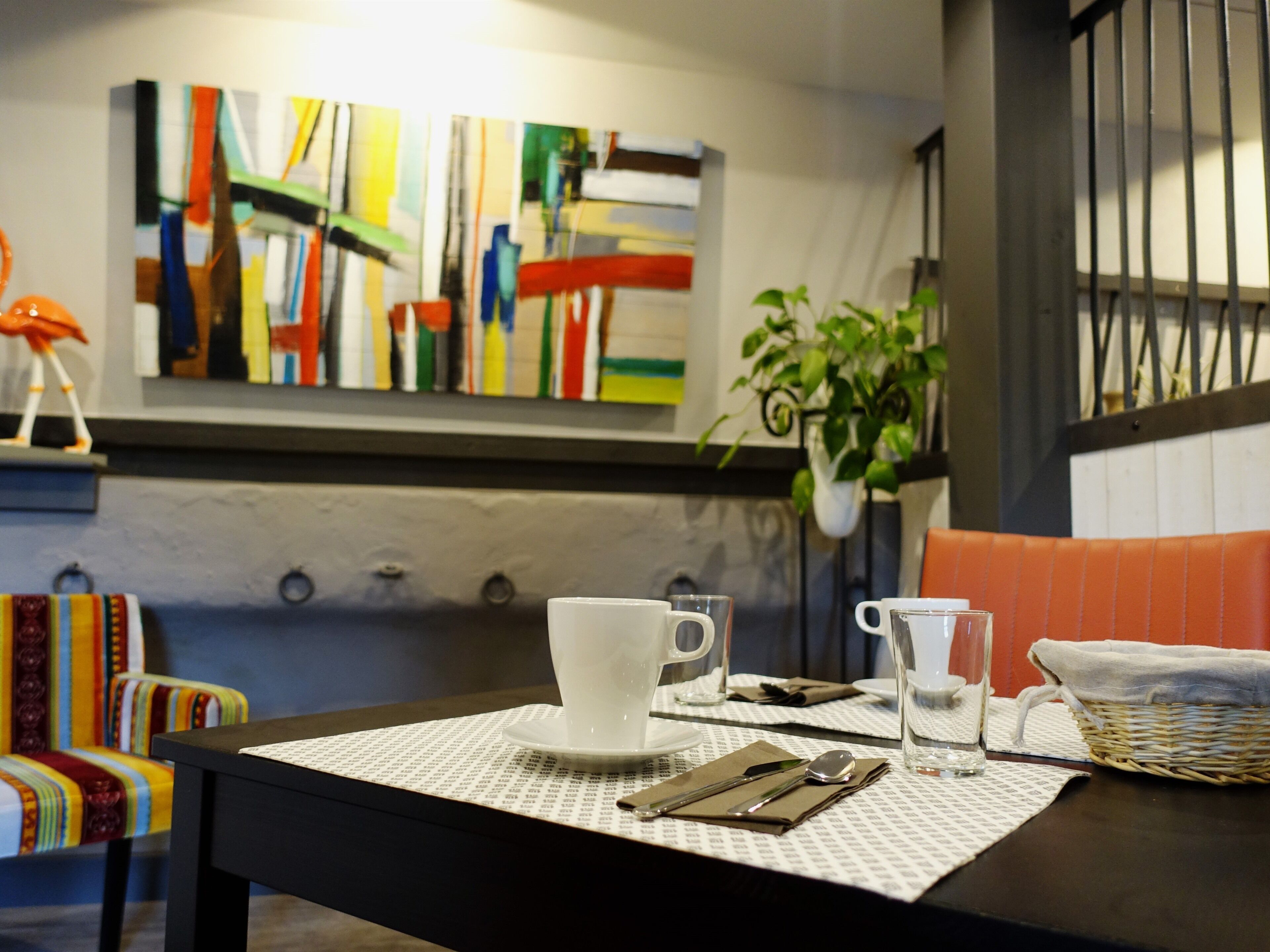 Una mesa de comedor con una servilleta blanca, una taza blanca, una copa, una cuchara y un cuchillo. Hay una silla colorida con un cojín a rayas y una planta en maceta en la pared.