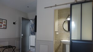 Habitació d'hotel amb un llit, una taula de nit, un bany amb un lavabo i un mirall, i una porta que condueix a una altra habitació.