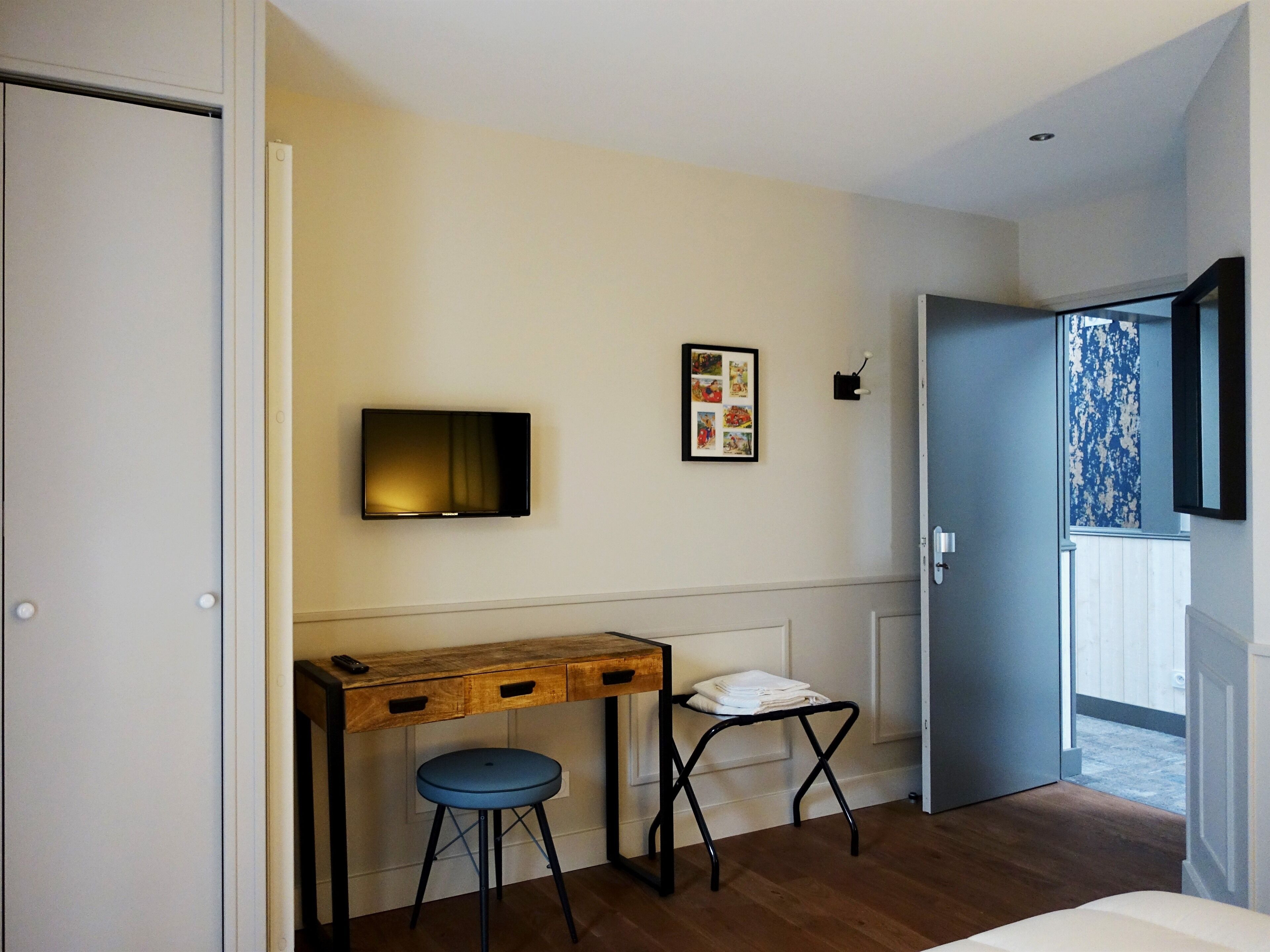 A room with a wooden desk, a blue stool, a TV mounted on the wall, and a door leading to a balcony.