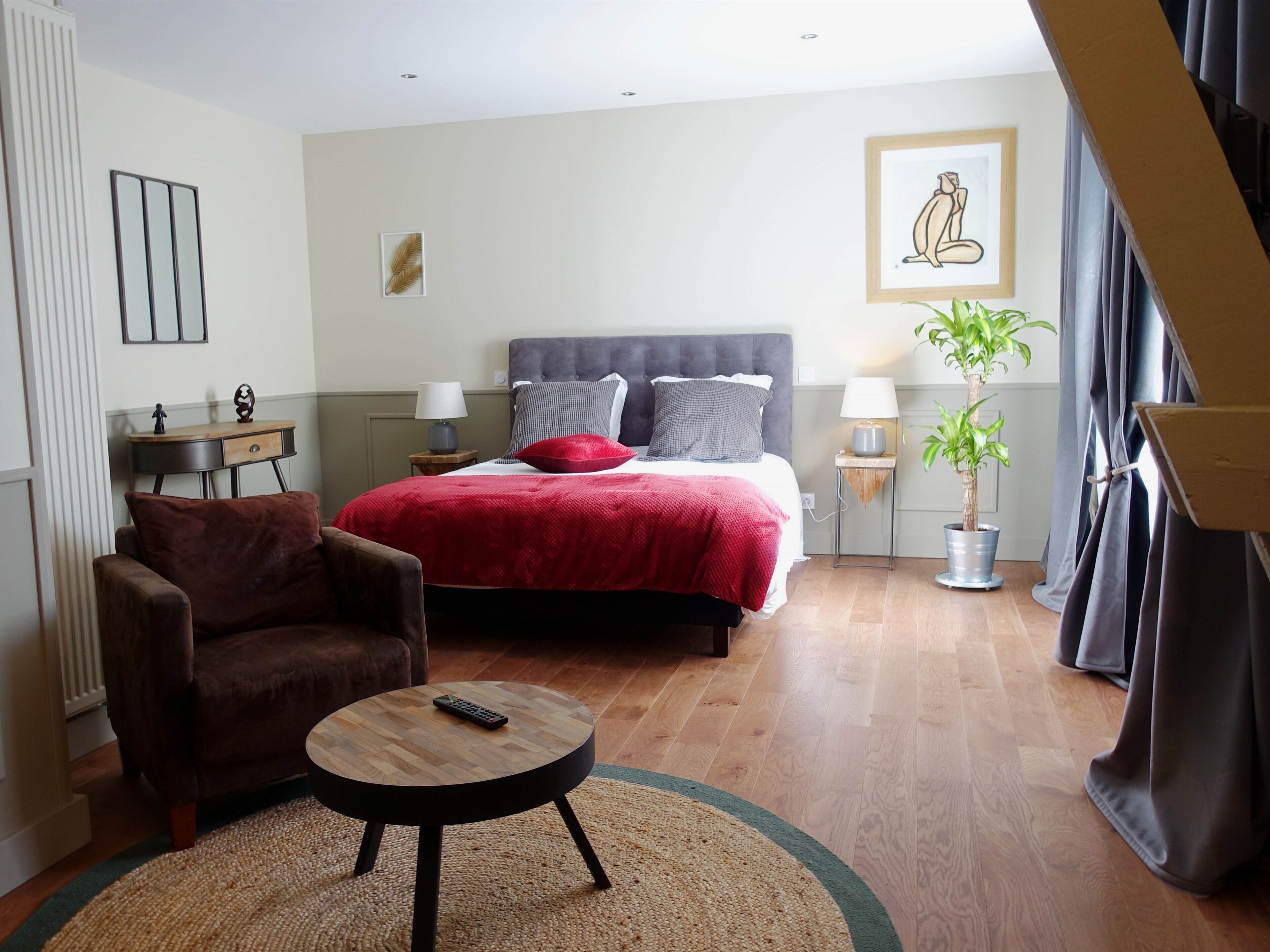 A bedroom with a bed, a chair, a small table, a lamp, a plant, and a painting on the wall.