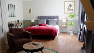 Une chambre à coucher avec un lit, une chaise, une petite table, une lampe, une plante et un tableau accroché au mur.