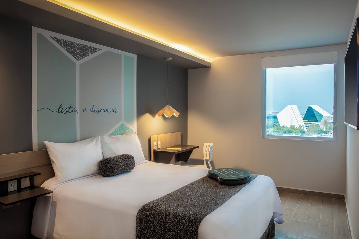 A modern hotel room with a bed, bedside table, lamp, and a window with a city view.
