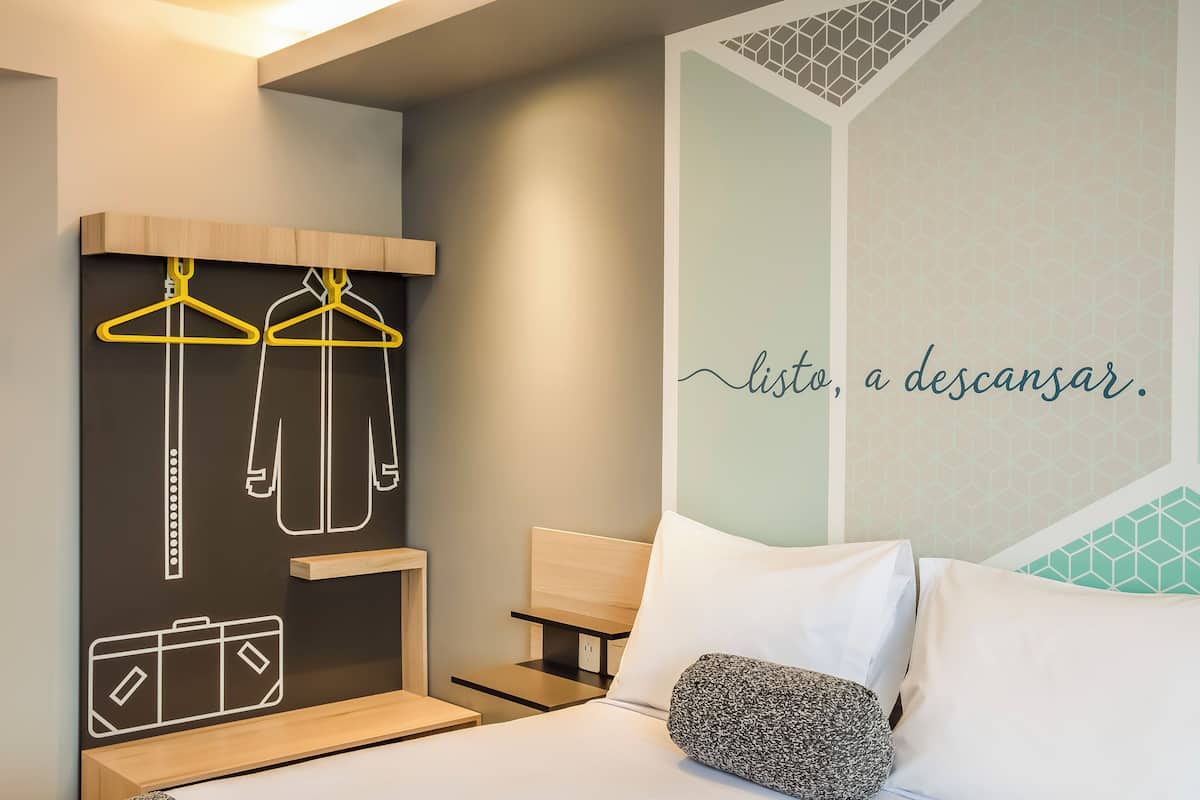 A hotel room with a bed, a nightstand, a wall-mounted coat rack, and a wall with a geometric pattern and text.