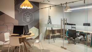 A modern office space with a glass partition, a desk with two monitors, a standing desk, and a lounge area with chairs.