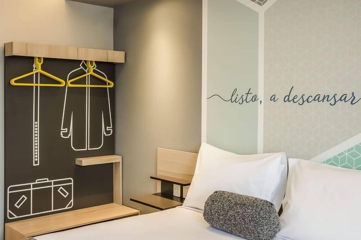 A hotel room with a bed, a bedside table, a wall-mounted coat rack with hangers, and a decorative wall with a geometric pattern and text.