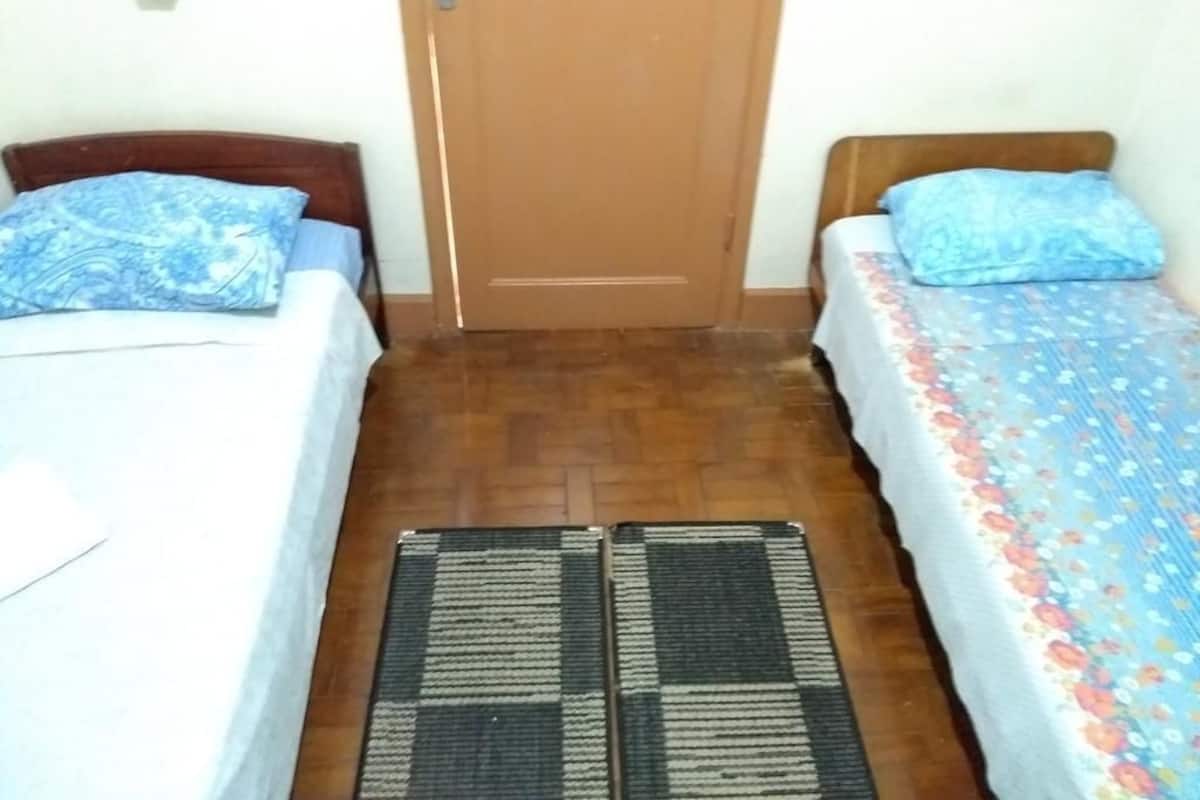 A room with two beds, each with a patterned bedspread and matching pillows. A wooden door is centered between the beds, and a small rug is placed on the floor.