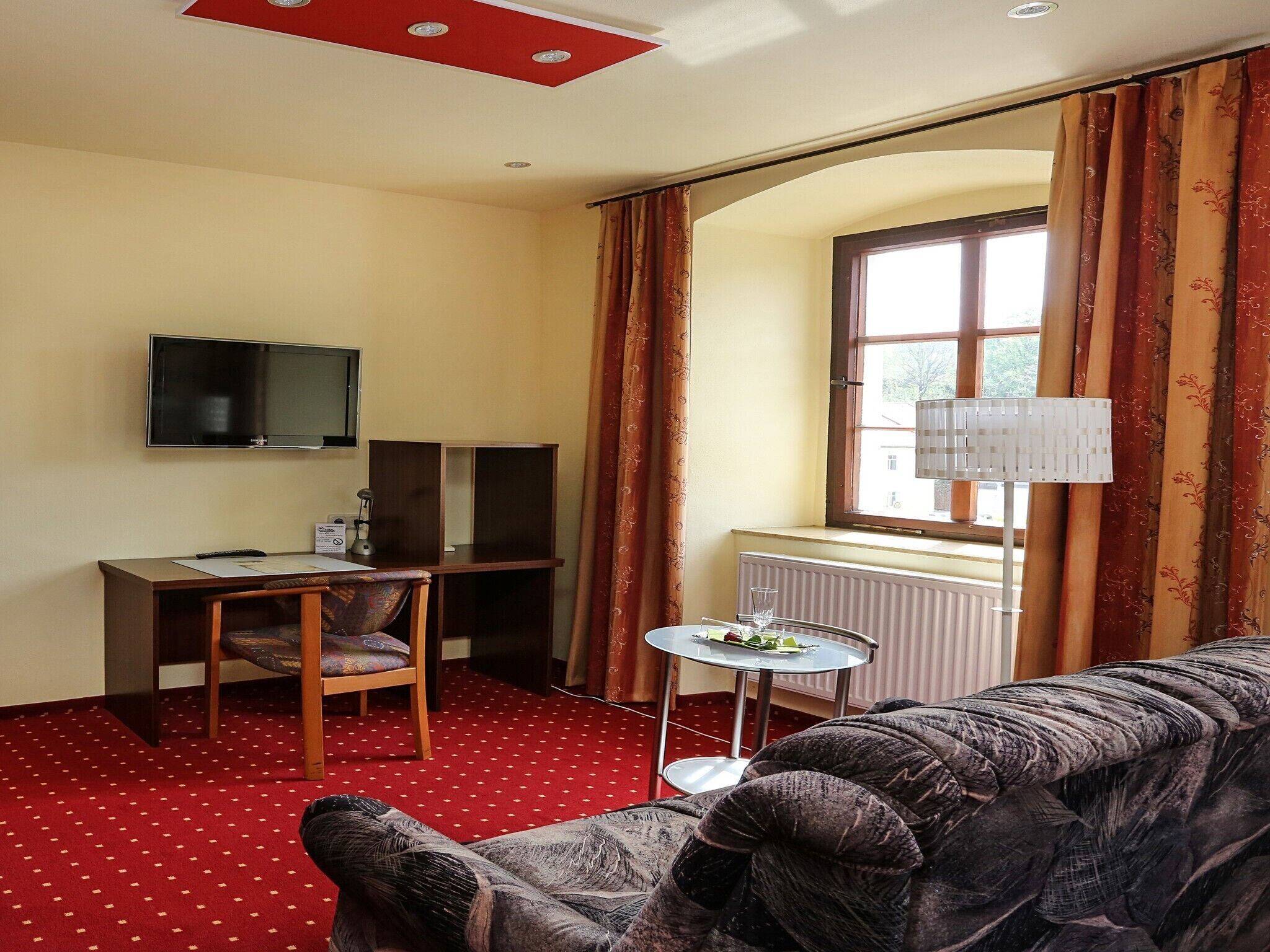 A hotel room with a red carpet, a sofa, a TV, a desk with speakers, a window with curtains, and a radiator.
