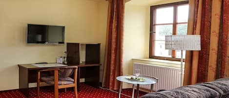 A hotel room with a red carpet, a sofa, a TV, a desk with speakers, a window with curtains, and a radiator.