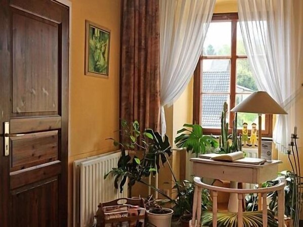 Una habitación con puerta de madera, un radiador, una ventana con cortinas, una mesa con lámpara y varias plantas en macetas.