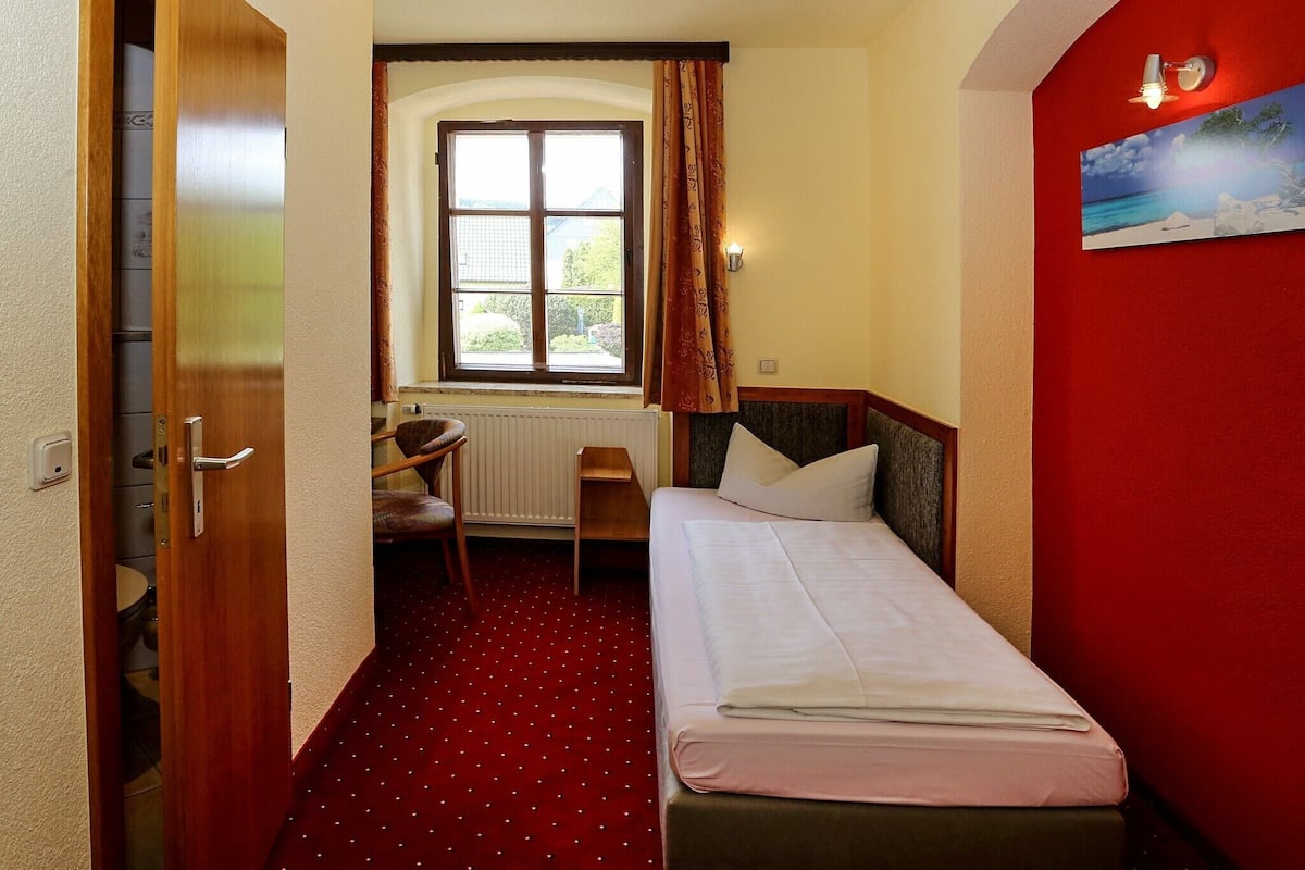 A hotel room with a bed, a small desk, a chair, a window with curtains, and a red accent wall.