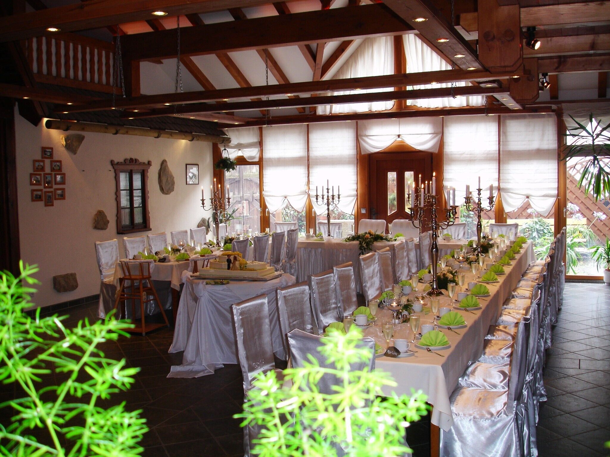 A long dining table set with white tablecloths, chairs, and candle holders, in a room with wooden beams and large windows.