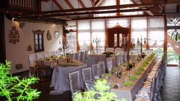 A long dining table set with white tablecloths, chairs, and candle holders, in a room with wooden beams and large windows.