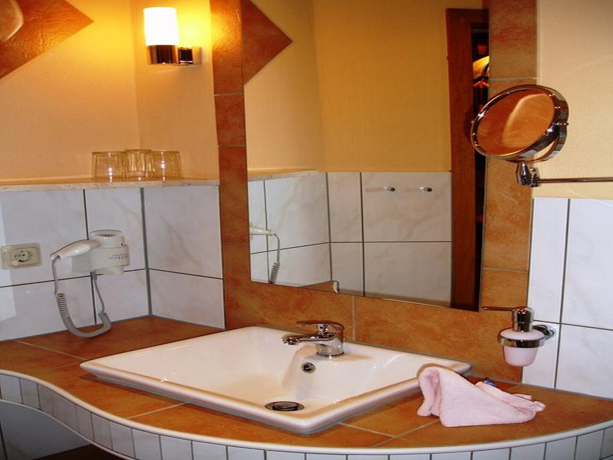 A bathroom with a white sink, a handheld hair dryer, a towel, a mirror, and a wall-mounted light fixture.