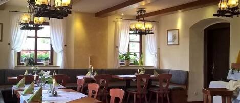 En restaurant med træborde og stole, lysekroner og store vinduer med gardiner.