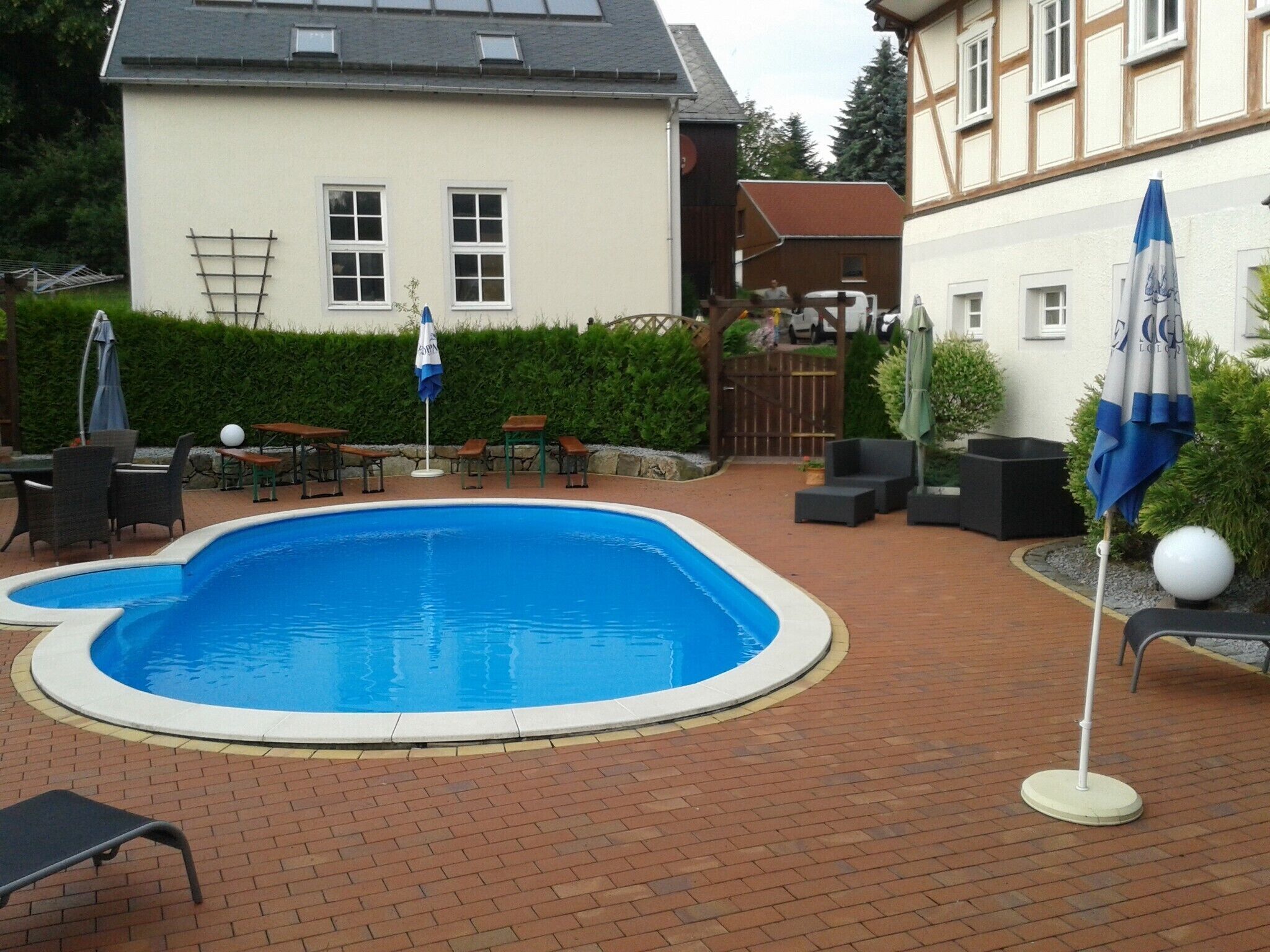 A swimming pool with lounge chairs and umbrellas, surrounded by a brick patio and a house in the background.