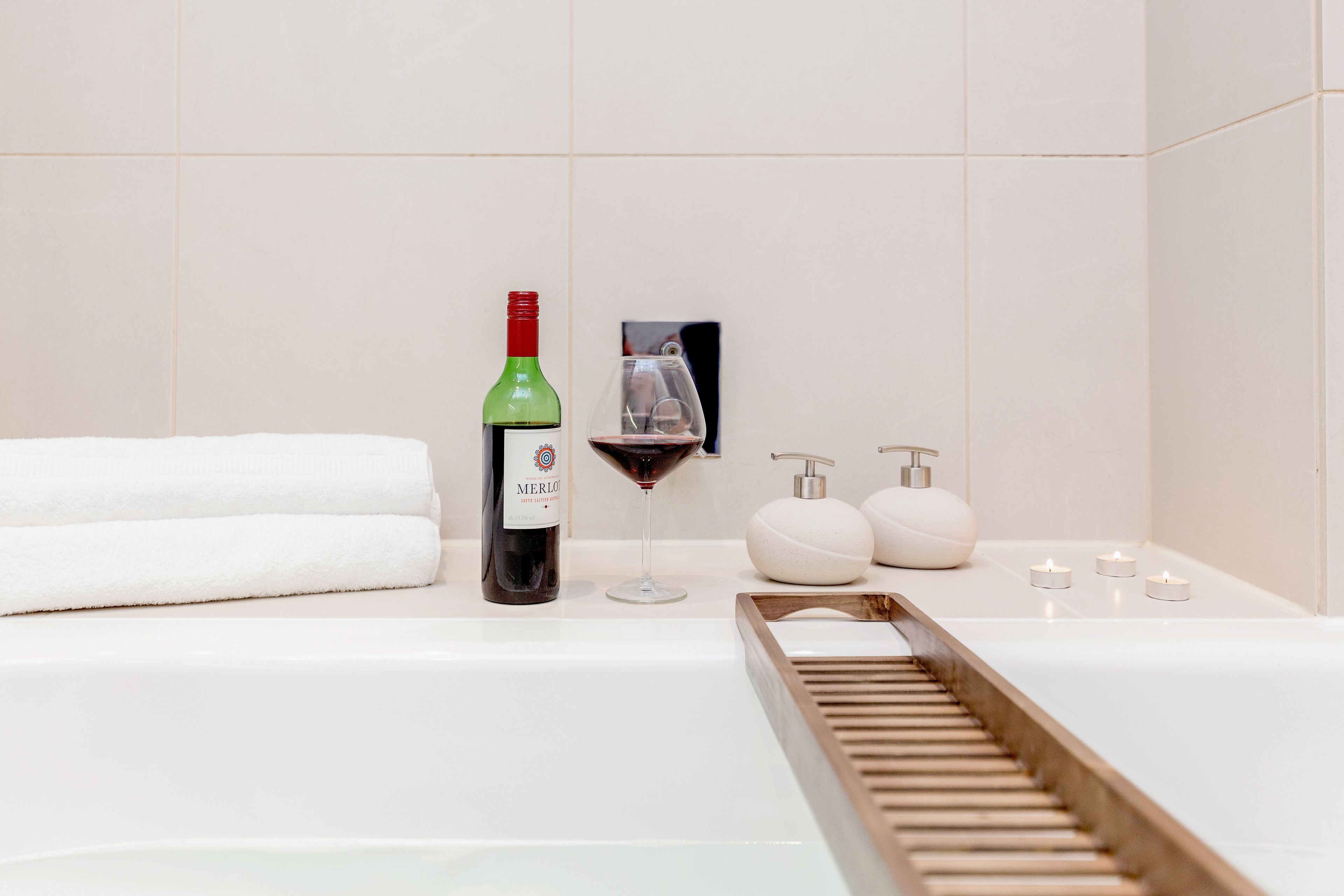 A bathroom with a bathtub, a bottle of Merlot wine, a wine glass, and toiletries.