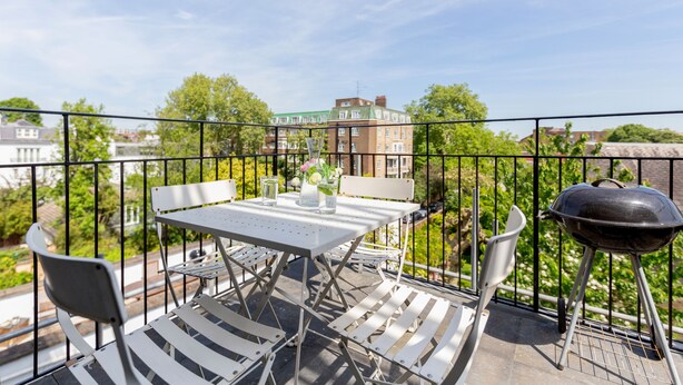 A balcony with a table and chairs, a grill, and a view of buildings and trees. - Kensington Apartment with terrace (London)