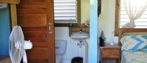 A small, compact room with a wooden door, a white fan, a toilet, a sink, and a bed with a striped bedspread.