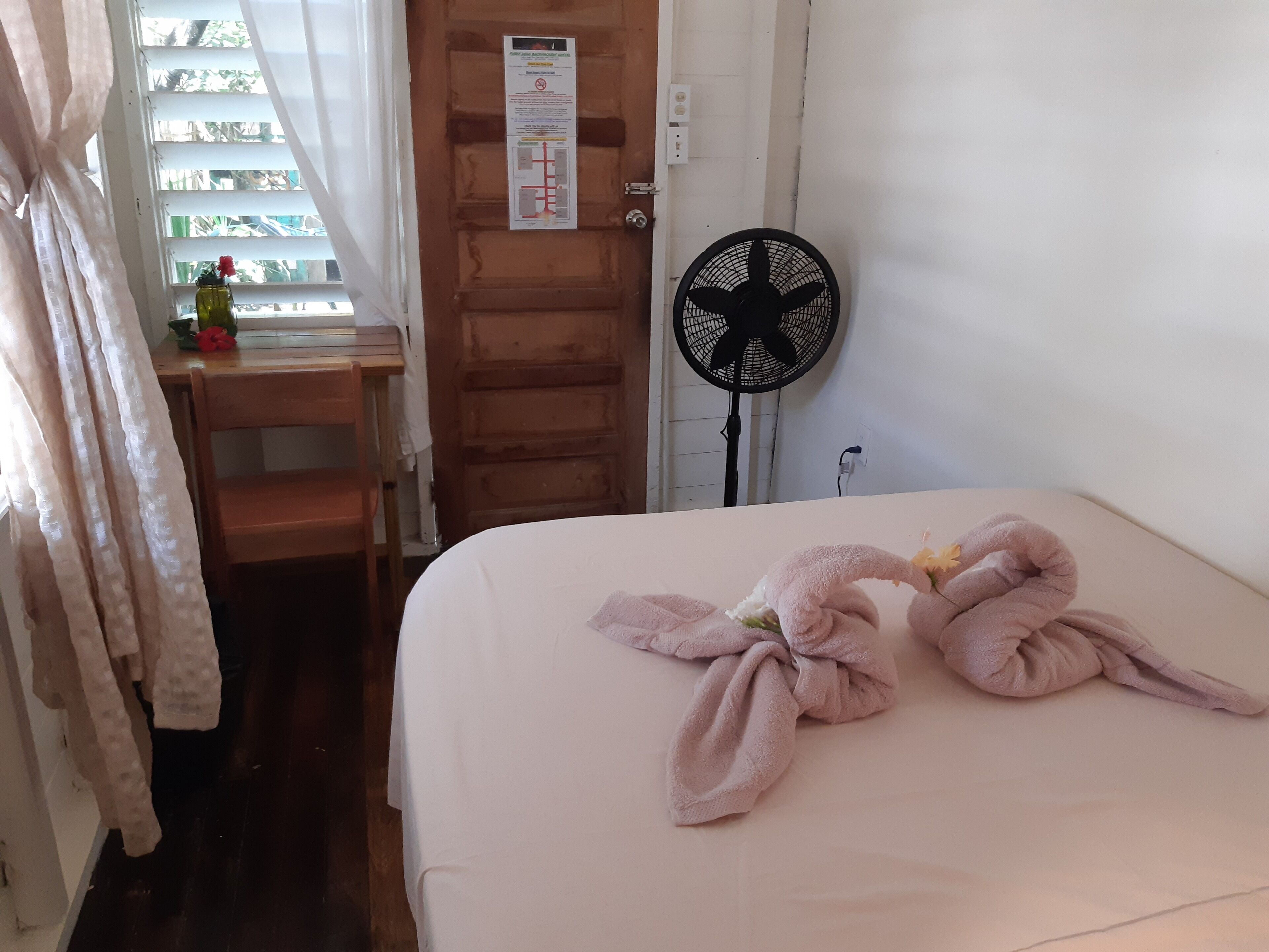 A neatly made bed with a towel arrangement, a wooden desk with a chair, a window with curtains, and a wall-mounted fan.