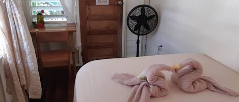 A neatly made bed with a towel arrangement, a wooden desk with a chair, a window with curtains, and a wall-mounted fan.