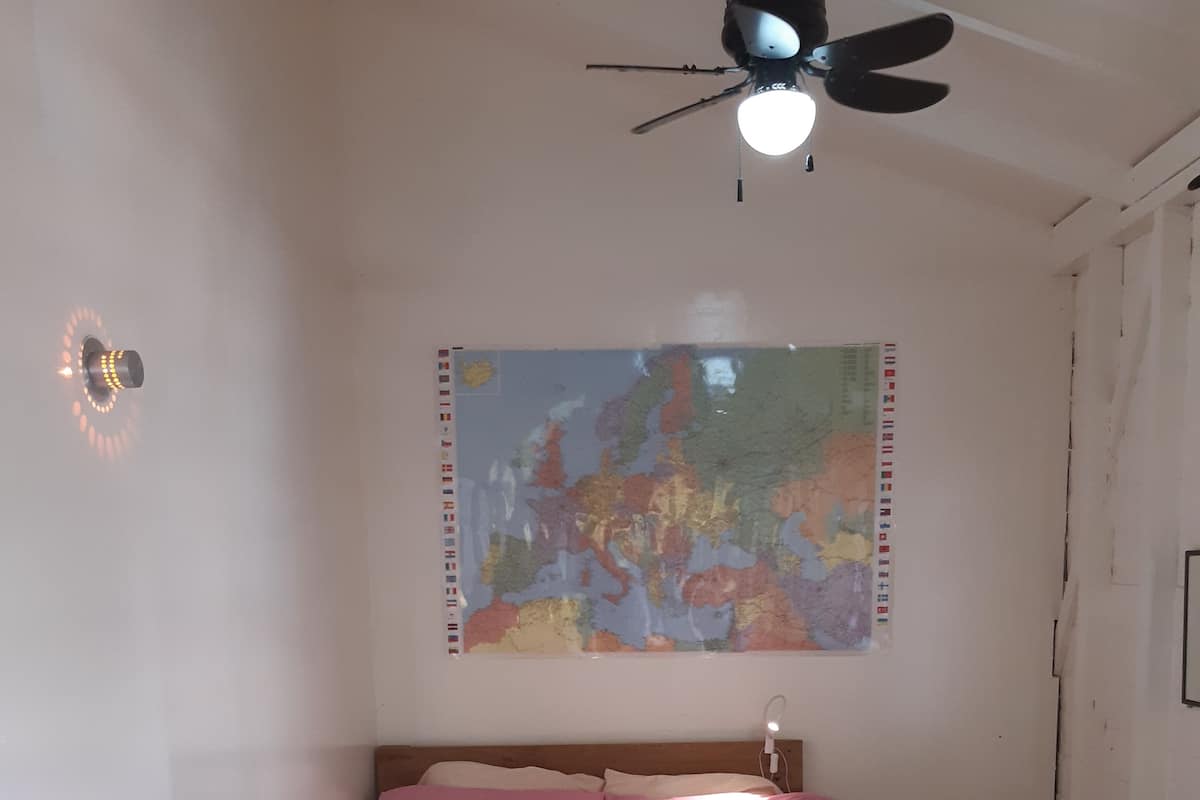 Un lit bien fait, avec une literie rose et blanche, une carte du monde accrochée au mur et un ventilateur de plafond.