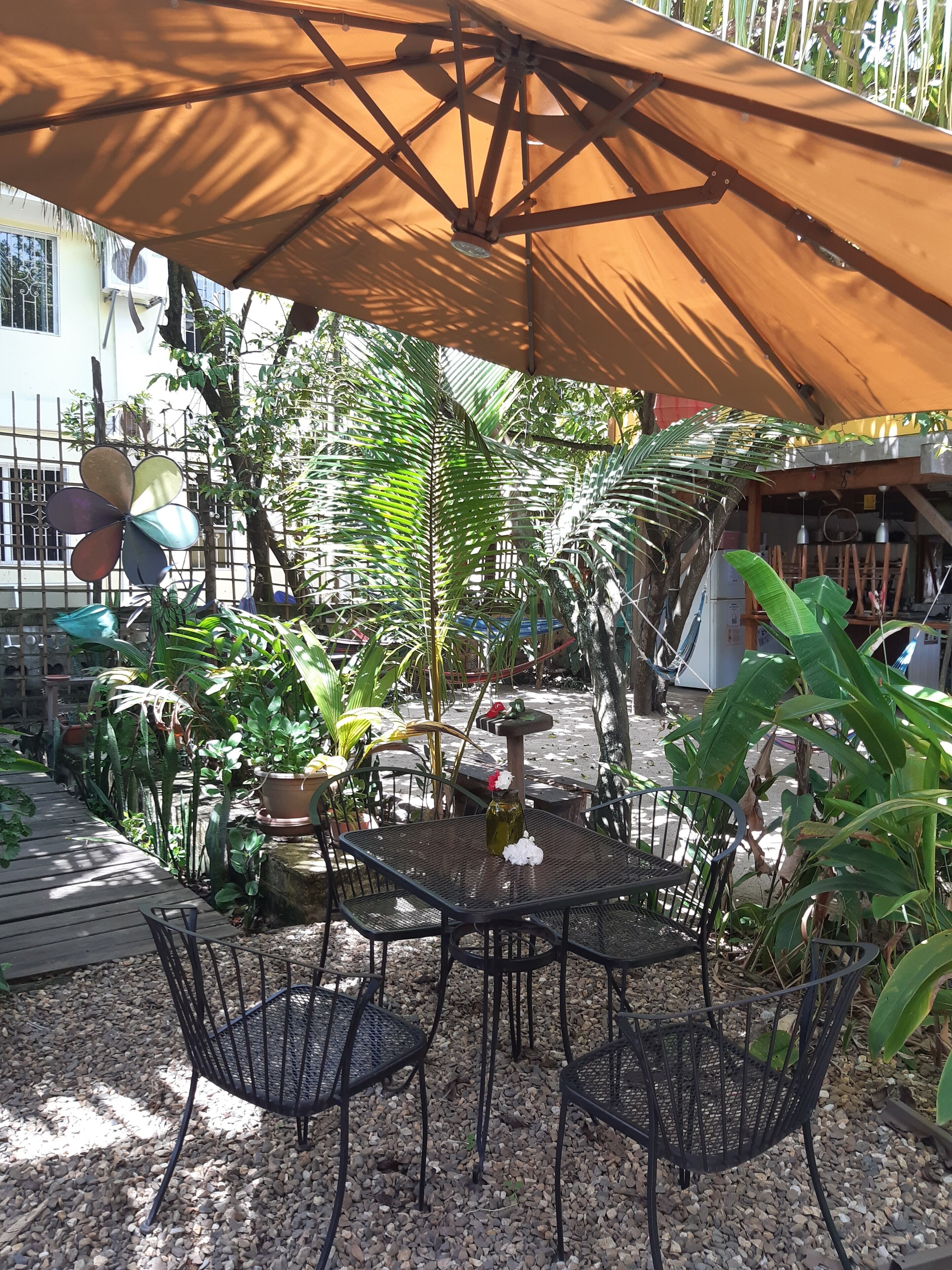 An outdoor patio area with a table and chairs under a large umbrella, surrounded by various potted plants and a decorative birdhouse.