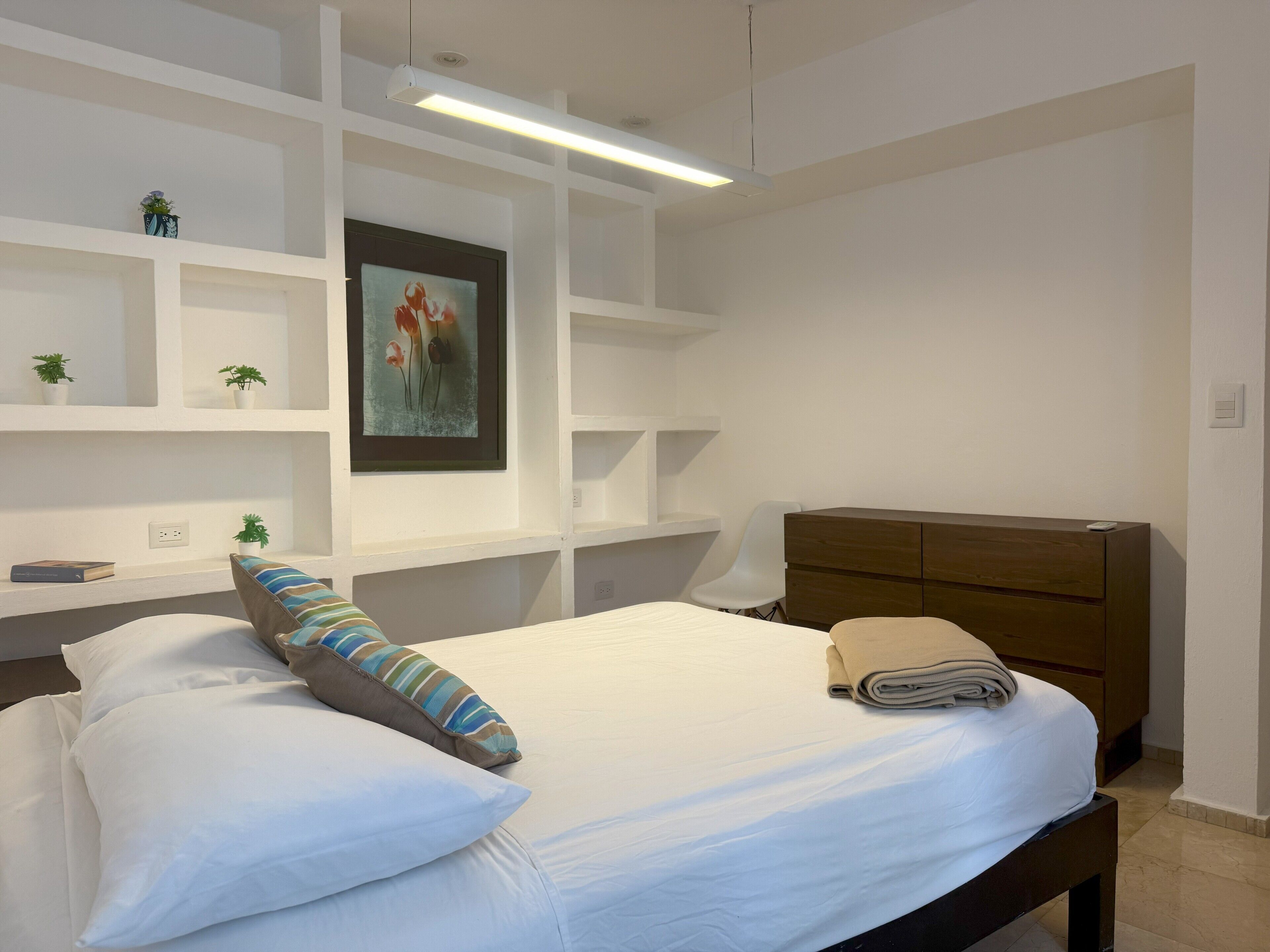 Un dormitorio con una cama, un aparador de madera, estanterías blancas y una obra de arte enmarcada en la pared.