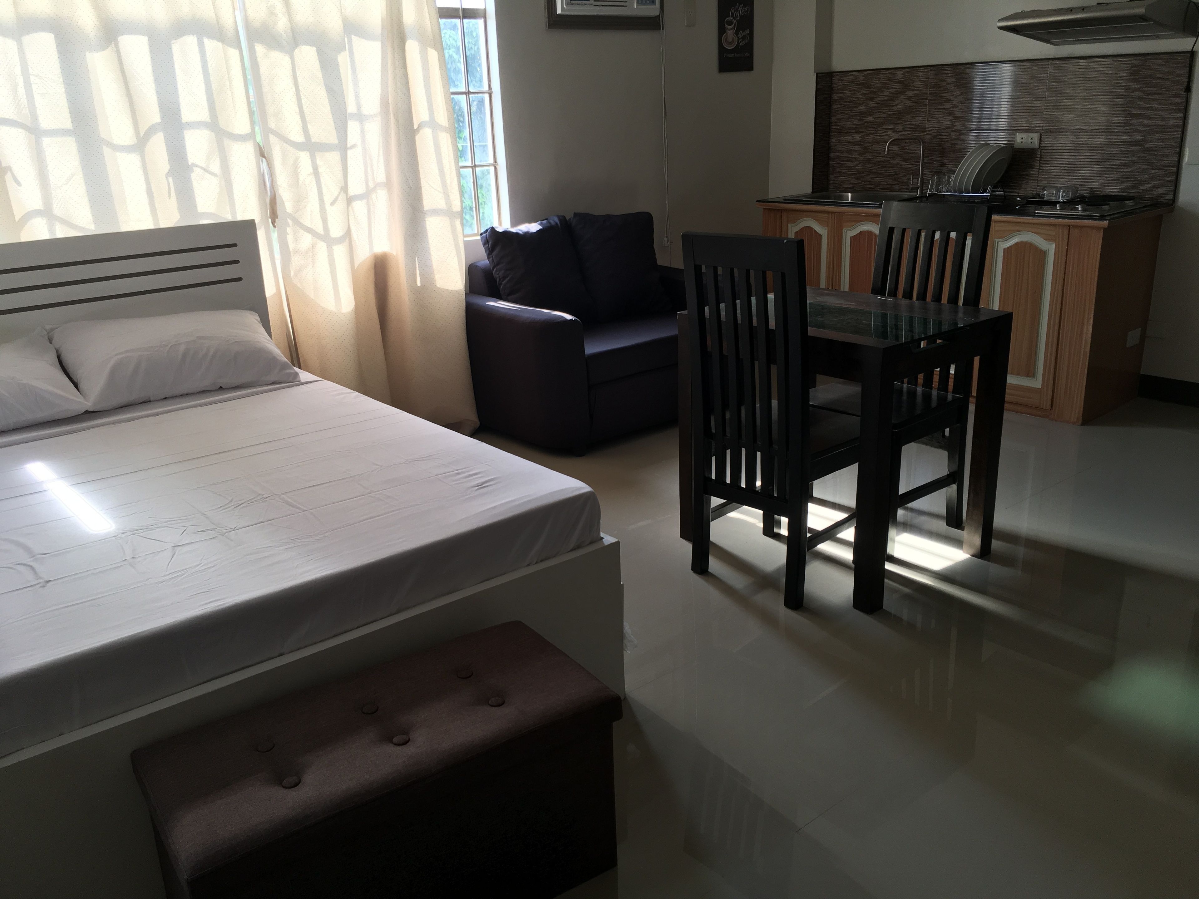 Un dormitorio con cama, mesita de noche, una silla, un sofá y una kitchenette al fondo.