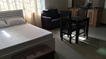 A bedroom with a bed, a nightstand, a chair, a sofa, and a kitchenette in the background.