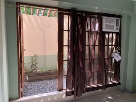 Una habitación con puerta de vidrio corrediza que da a un balcón, una ventana con cortina y un equipo de aire acondicionado en la pared.
