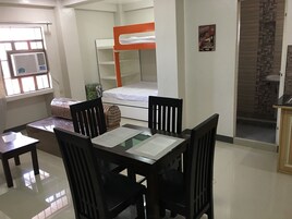 Habitación pequeña con literas, una mesita con sillas, ventana con aire acondicionado y suelo de baldosas.