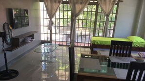 Una habitación con una mesa de vidrio, sillas de madera, una cama con ropa de cama verde, un televisor en la pared y una ventana con cortinas.
