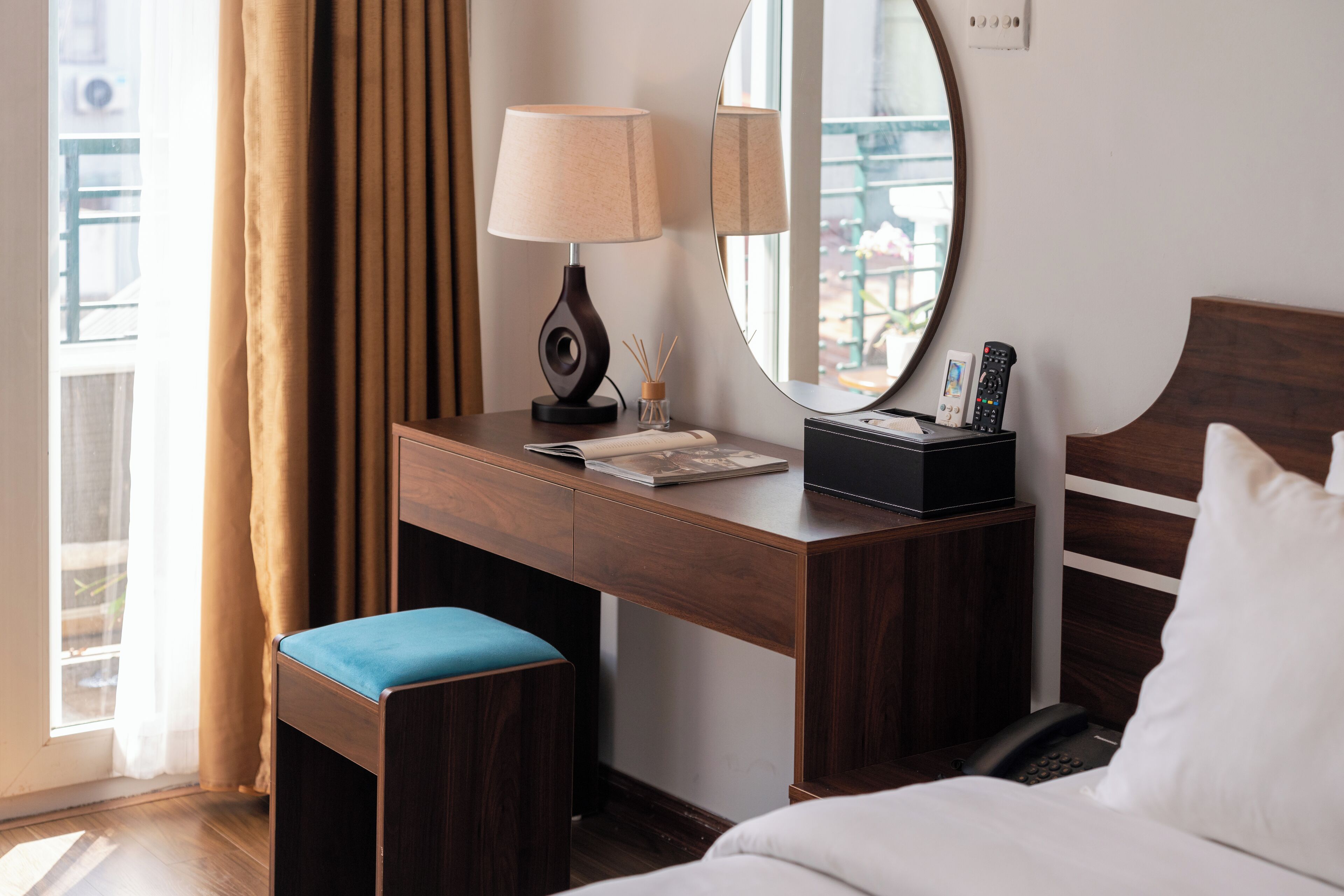 A hotel room with a wooden desk, a lamp, a mirror, a bedside table with a phone, and a bed with white bedding.