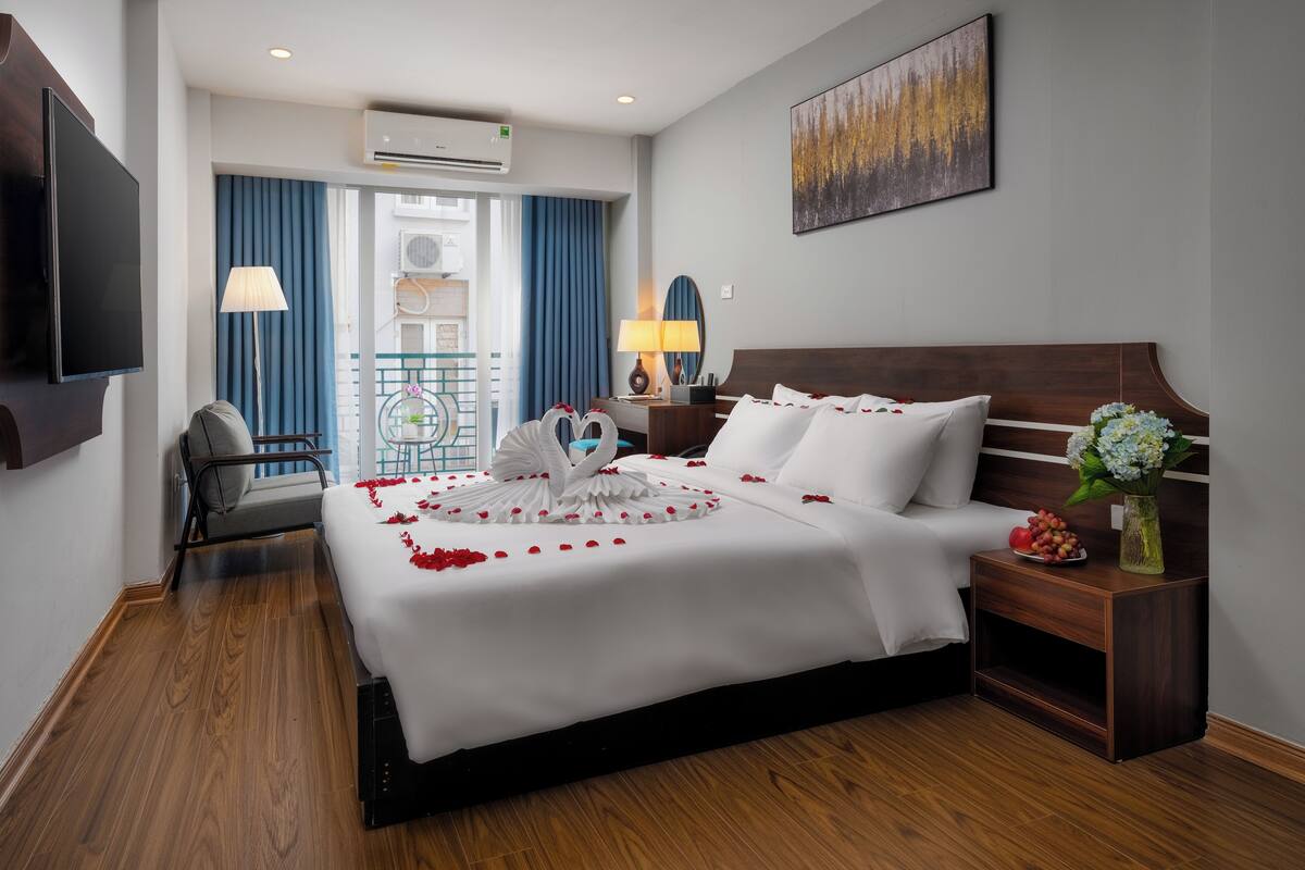 Une chambre d’hôtel avec un grand lit, une télévision, un bureau, une chaise, un balcon avec des rideaux bleus et un tableau au mur.
