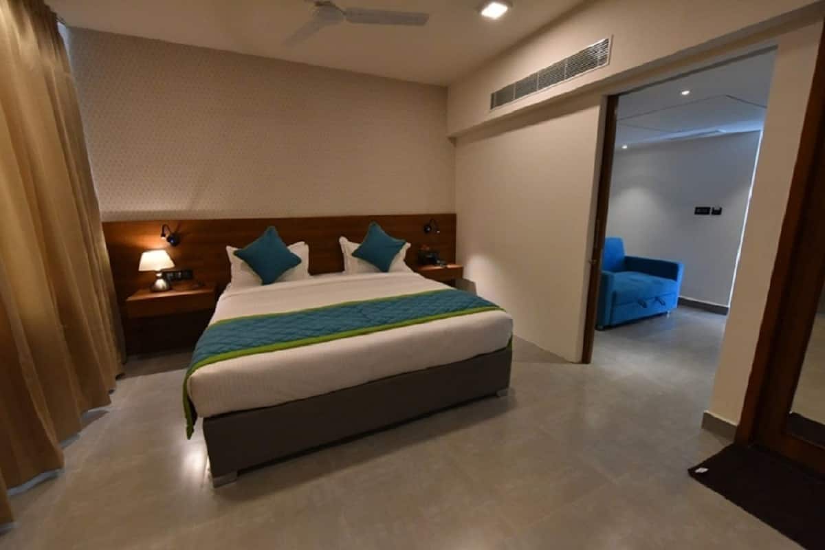 A hotel room with a large bed, a nightstand with a lamp, a blue chair, and a door leading to another room.