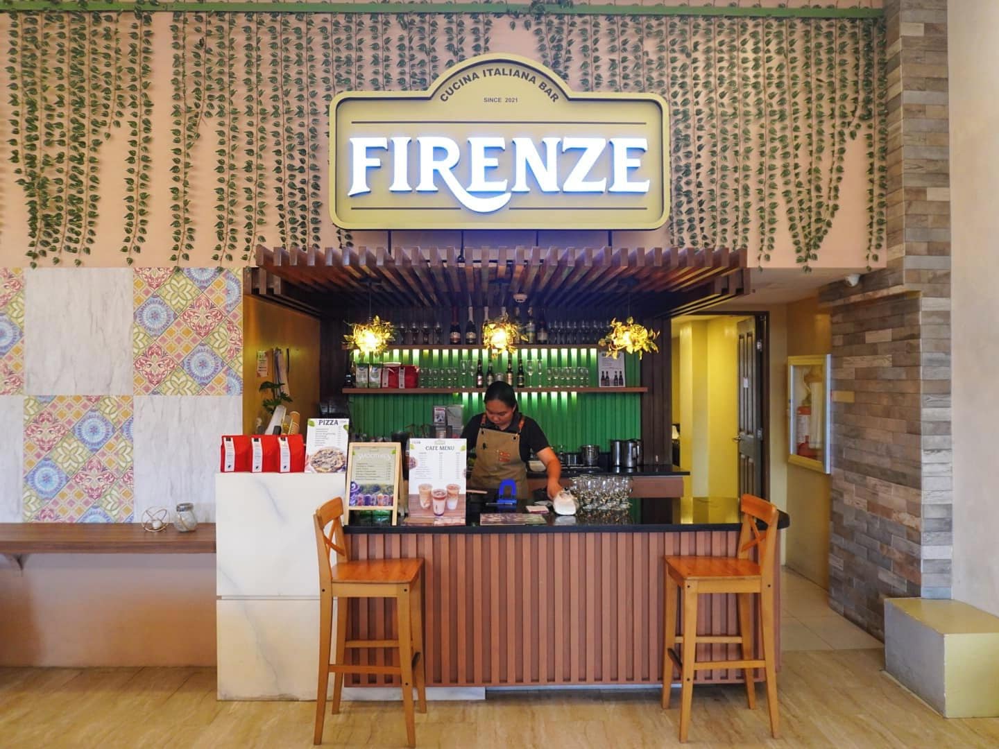 A bar named 'FIRENZE' with a bartender, a menu board, and a counter with two stools.