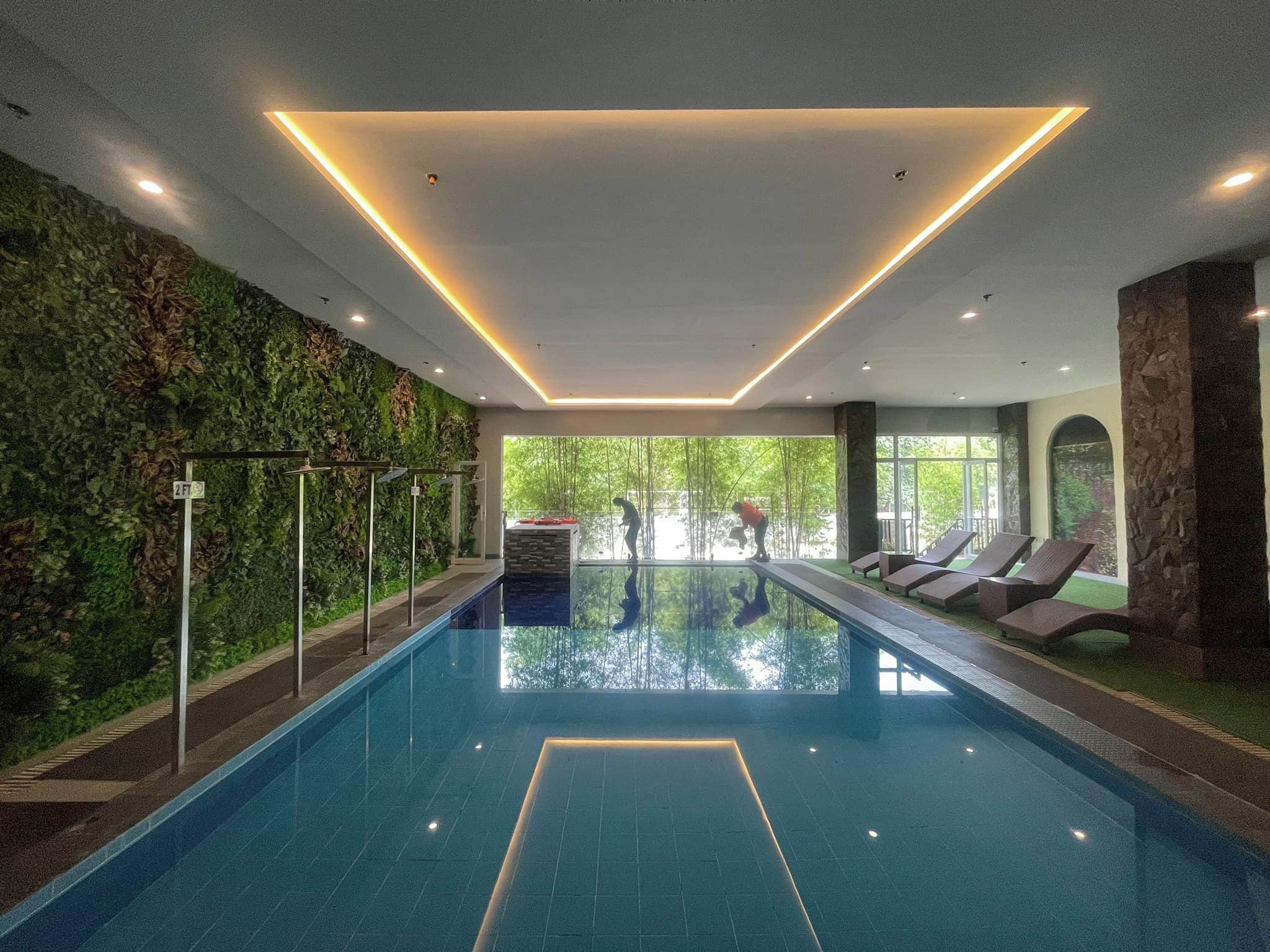An indoor swimming pool with a clear view of the outdoors, surrounded by greenery and modern lighting.