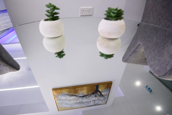 Un interior moderno con una mesa de vidrio reflectante, dos plantas en macetas, una obra de arte enmarcada y un enchufe en la pared.