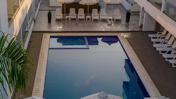 Un área de piscina en la azotea con sillones de descanso y sombrillas, rodeada de palmeras y edificios modernos.