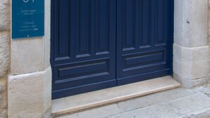 Une porte à double battant de couleur bleu foncé, surmontée d’une arche décorative en métal, est intégrée à un bâtiment en pierre. Une enseigne sur le côté gauche indique : « SOTRANI TEMPORARY HOUSE, 1er étage, T+33 847 1684, www.sotrani.it ».