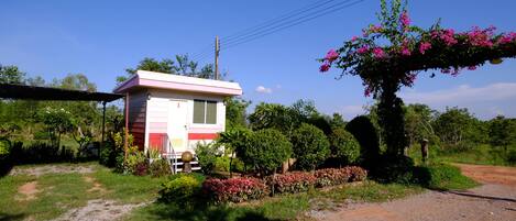 A small, single-story building with a pink and white exterior, surrounded by lush greenery and a gravel path.