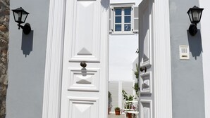 Una puerta blanca con una corona decorativa, flanqueada por dos faroles en la pared, que da paso a un patio cubierto con plantas en macetas.