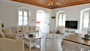Amplia sala de estar con sofá, mesa de centro, mesa de comedor y sillas. Un televisor está montado en la pared, y hay una puerta que da a otra habitación.