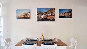 Un comedor con una mesa de madera dispuesta para cuatro personas, sillas blancas y tres cuadros de temática costera en la pared.