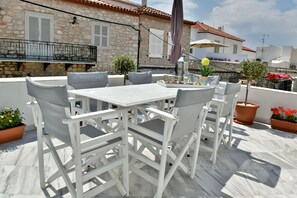 Una terraza con una mesa y sillas blancas, plantas en macetas y un edificio de piedra al fondo.