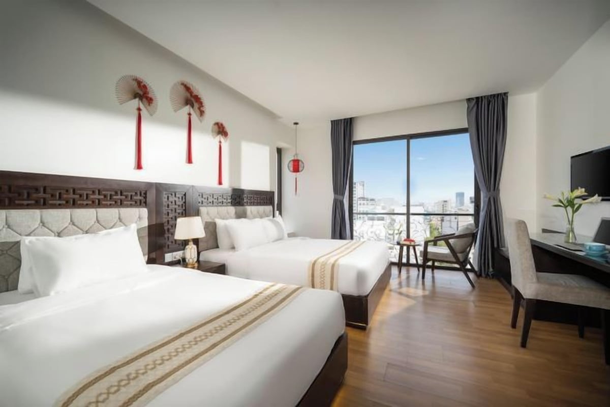 A hotel room with two beds, a desk, a chair, and a large window offering a city view.